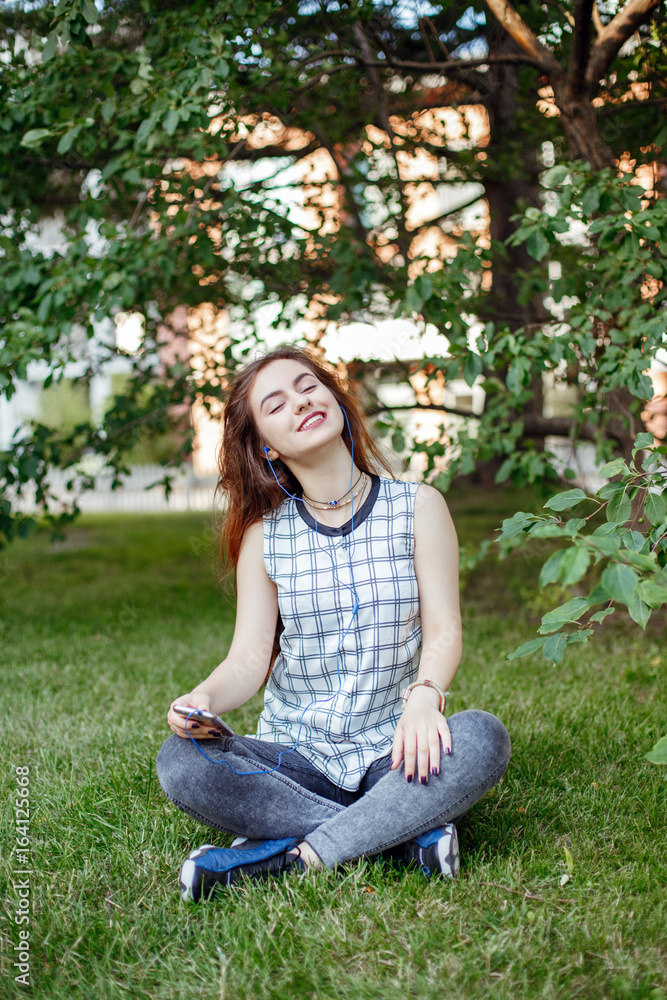 Portrait of beautiful white Caucasian woman with long red hair in jeans and plaid shirt sitting on grass outside in park. Woman with earbuds earphones listening to music on her phone on Internet. Portrait of beautiful white Caucasian woman with long red hair in jeans and plaid shirt sitting on grass outside in park. Woman with earbuds earphones listening to music on her phone on Internet.