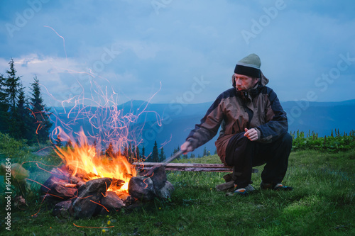 Active hiker sitting near camping fire at twilight in the mountain