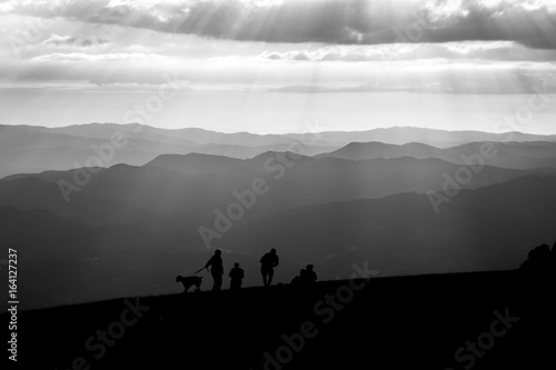 Some people and a dog on top of a mountain  with other mountains and hills in the background  and sunrays coming out of the clouds