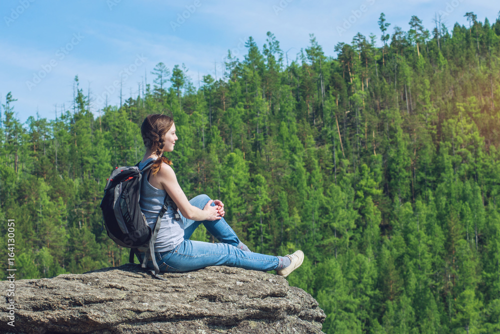Girl with backpack sitting on a rock on top of a mountain on the background of green forest