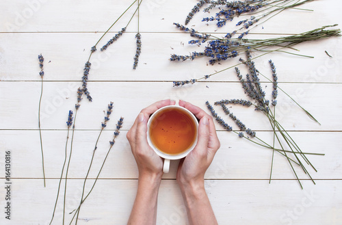 A cup of tea in female hands and lavender on a white wooden background. Top view.