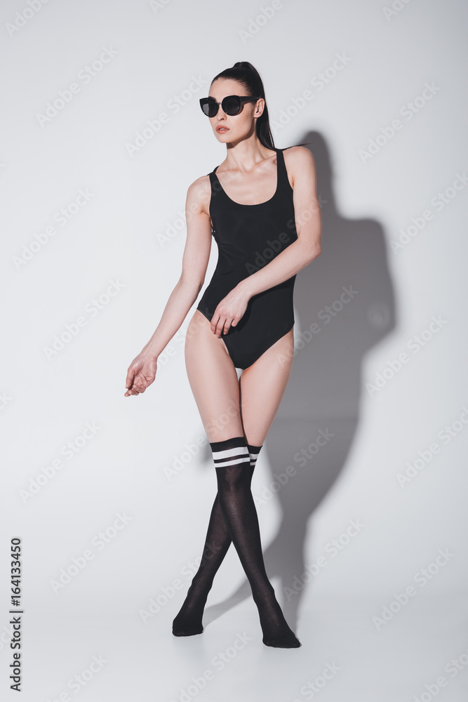 Attractive young brunette woman in fashionable leotard and sunglasses posing in studio