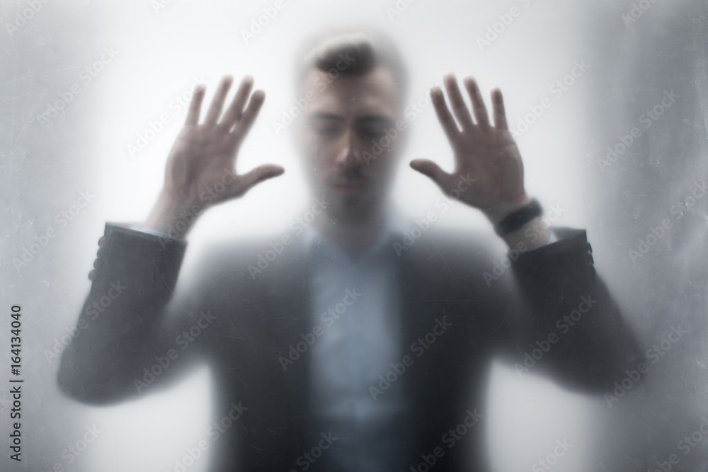 Silhouette of upset businessman with closed eyes touching glass with hands behind window