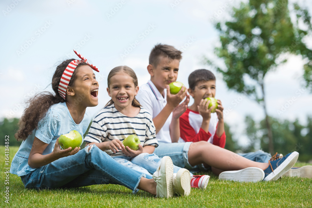Fototapeta premium Happy multiethnic children eating green apples while sitting together on green grass