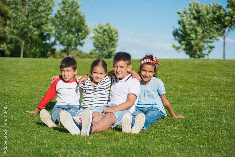 Adorable multiethnic kids sitting embracing in green grass and smiling at camera Adorable multiethnic kids sitting embracing in green grass and smiling at camera