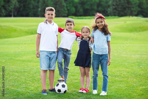 Adorable multiethnic kids standing with soccer ball and smiling at camera in park