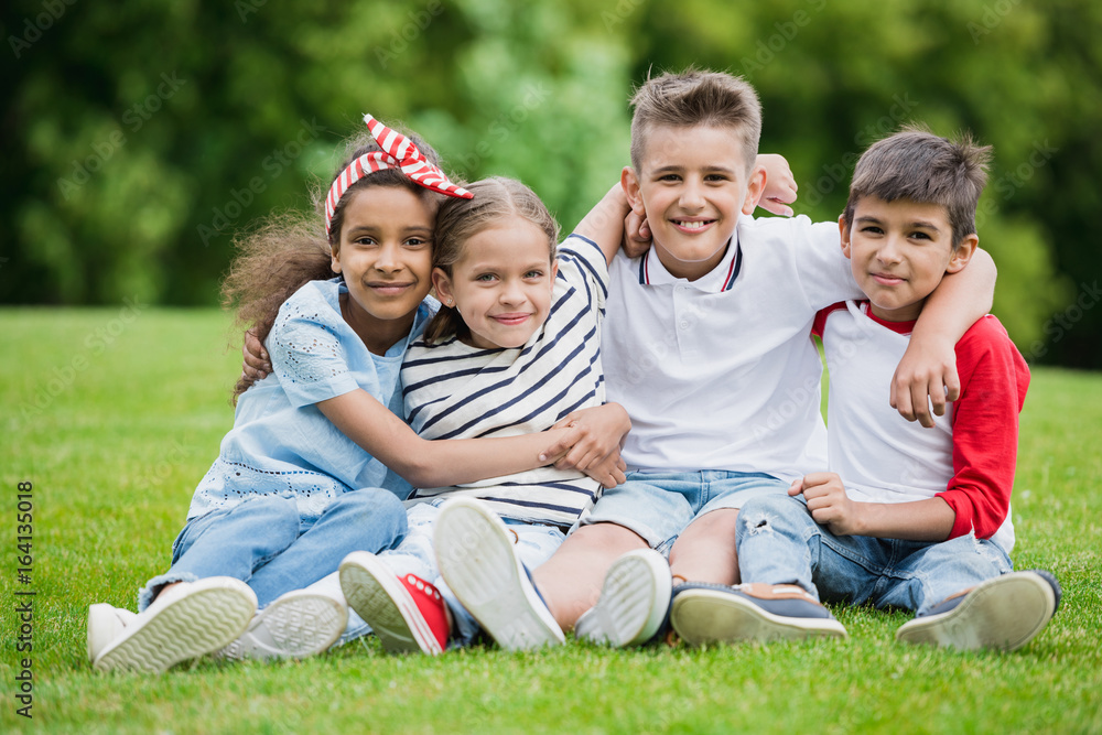 Happy multiethnic kids sitting embracing and smiling at camera in park Happy multiethnic kids sitting embracing and smiling at camera in park