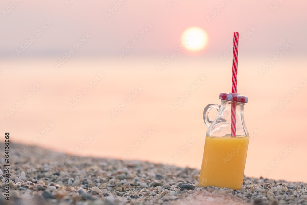 Glass bottle of fresh orange juice on beach at sunset over sea