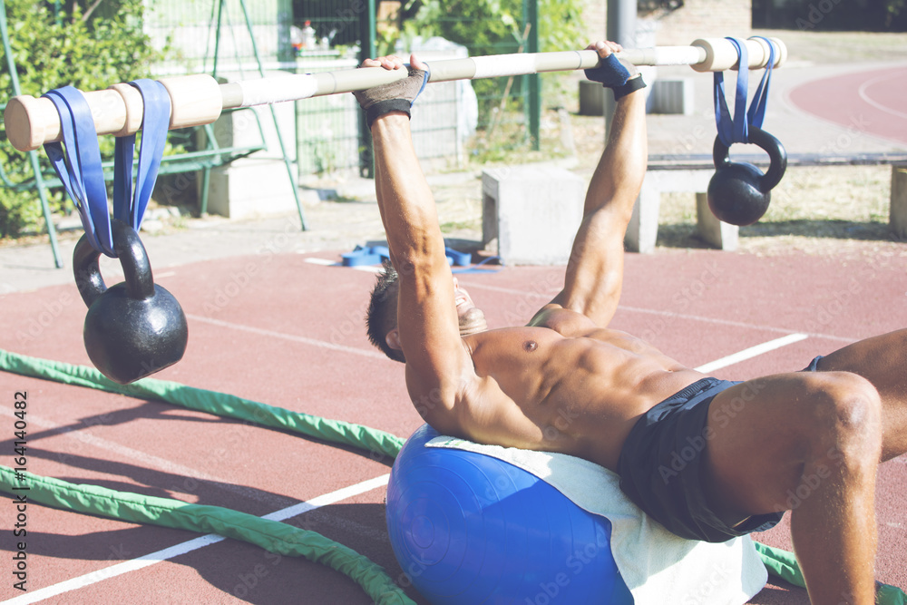 © Addoro - Handsome middle aged man working out on a running track. Healthy adult man doing push ups using a kettlebell and wooden barbell on the yoga ball. Tanned skin and shirtless middle-aged man.