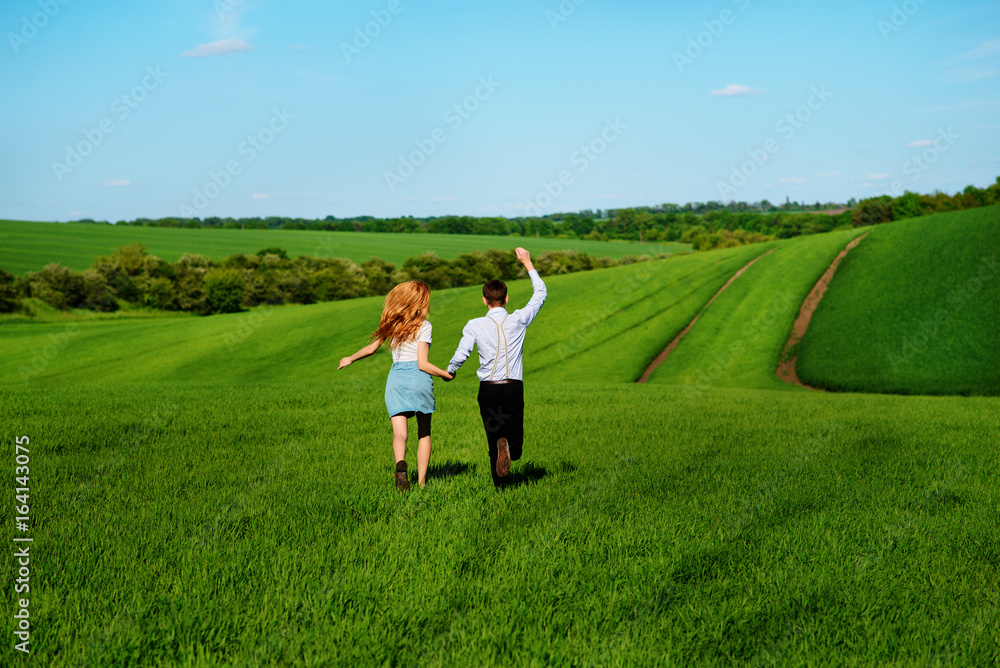 Young happy lovers running on meadow with green grass and blue sky, From the back Young happy lovers running on meadow with green grass and blue sky, From the back