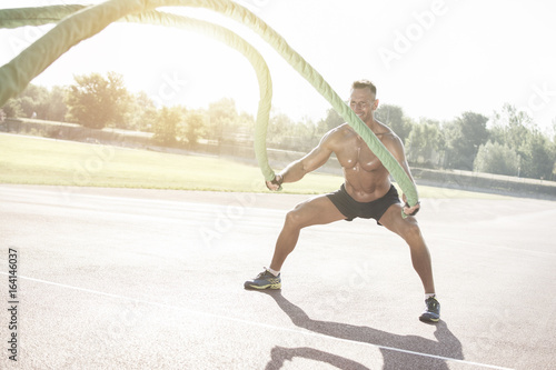 Handsome middle aged man working out on a running track. Healthy adult man doing battle rope exercise. Tanned skin and shirtless middle-aged man. 