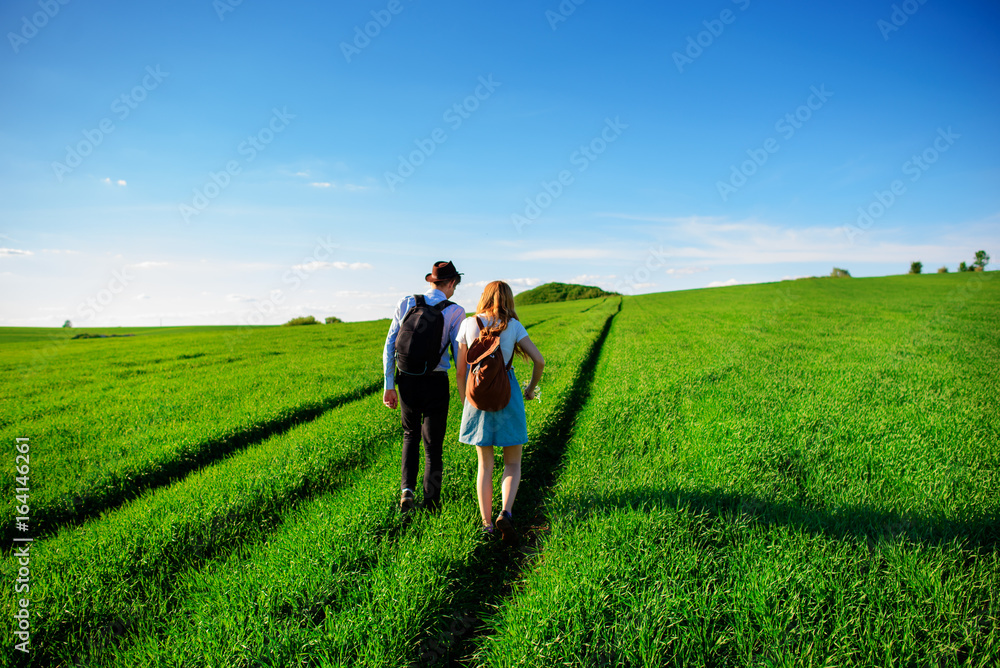 With a backpack, a man in a hat and a woman with long hair go along the path. A couple walks along the meadow With a backpack, a man in a hat and a woman with long hair go along the path. A couple walks along the meadow