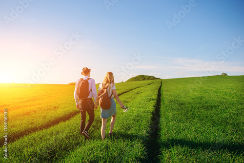 With a backpack, a man in a hat and a woman with long hair go along the path. A couple walks along the meadow