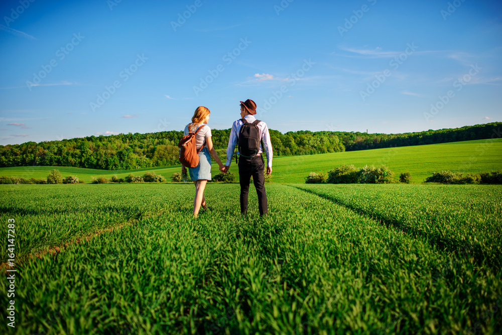 With a backpack, a man in a hat and a woman with long hair go along the path. A couple walks along the meadow With a backpack, a man in a hat and a woman with long hair go along the path. A couple walks along the meadow