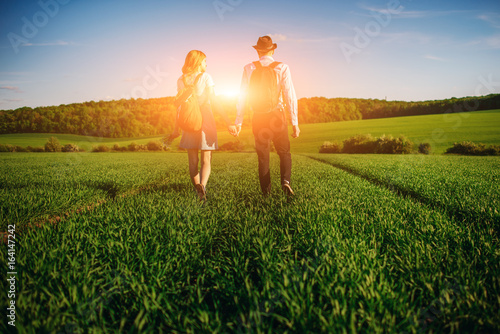 With a backpack, a man in a hat and a woman with long hair go along the path. A couple walks along the meadow