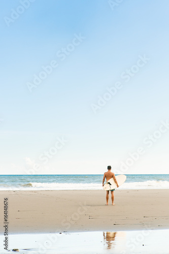 Handsome boy in blue swimming shorts and cap stand with long surf surfing board wait on surf spot at sea ocean beach. White blank surfboard. Concept of power, freedom, new modern life, generation Y.