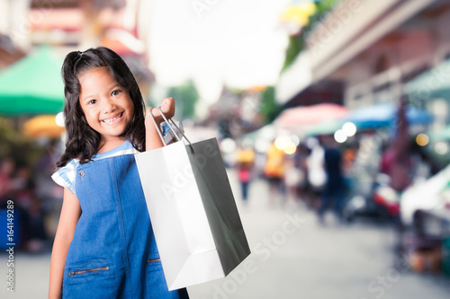 pretty little asian girl in blue jeans dungarees holding shopping bags with street market background.portrait of young kid smiling and looking at camera