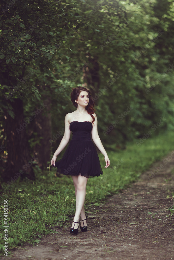 Beautiful young brunette girl in short black dress walking through the woods