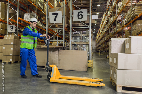 Male worker wearing blue uniform jacket and hardhat driving pallet truck towards pallets with boxes.