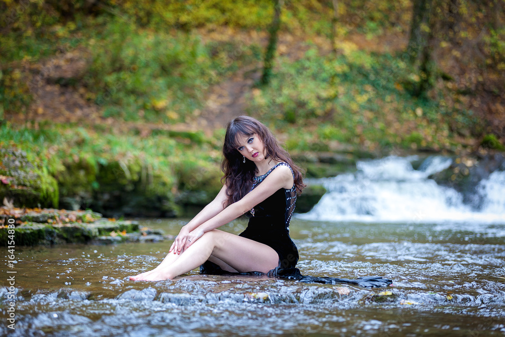 Cute brunette sitting in river raising legs and holding black dress Cute brunette sitting in river raising legs and holding black dress