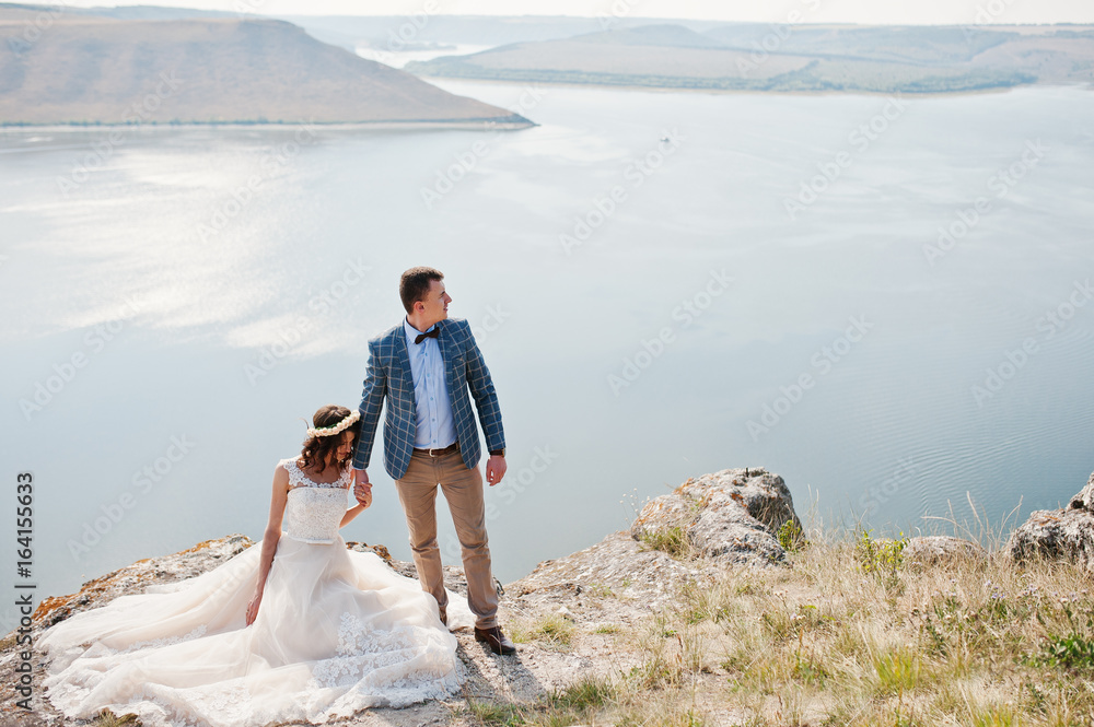 Gorgeous bride sitting on the stone while her husband stands next to her on the cliff with lake in the background.