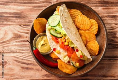 Tasty fish taco on plate with sauce and chips on wooden background