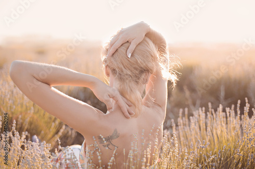 The neck and elegant tender hand of Young beautiful blonde Girl on lavender field in golden sunset light. Romantic Bare back woman take sun bath on floral field having vacations in Provence France