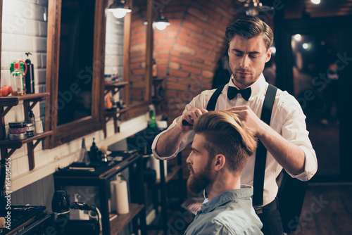 Young stylish bearded guy is in a barber shop, getting brend new haircut from a classy dressed stylist. Both are serious and attractive