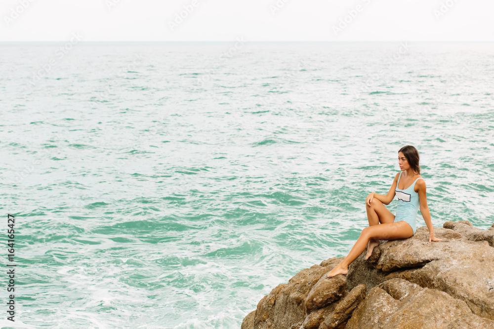 Pretty lady in full body swimsuit with long healthy hairs sit on big stone on the rock beach during sea ocean storm and touch her knee. Big waves near her legs. Concept of danger, sad, bad emotions. Pretty lady in full body swimsuit with long healthy hairs sit on big stone on the rock beach during sea ocean storm and touch her knee. Big waves near her legs. Concept of danger, sad, bad emotions.