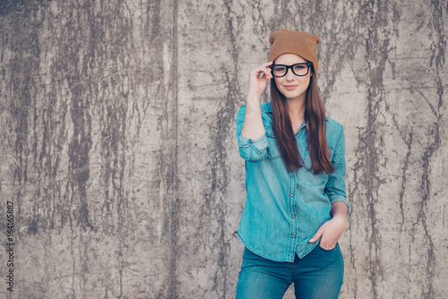 Hot attractive young girl is standing on the concrete wall`s background outdoors, fixing her glasses. She is wearing casual jeans outfit and brown hat