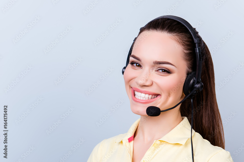 Successful and very cheerful young brunette girl. She is in a headset, standing on a pure light background. She is in a casual yellow tshirt. Has a beaming smile