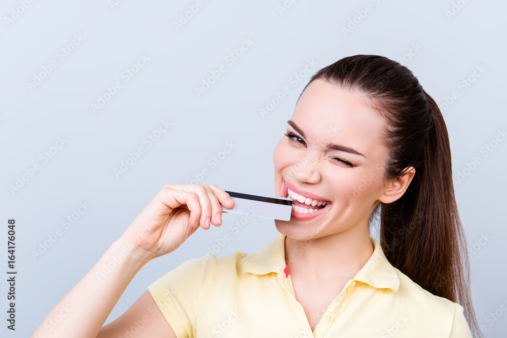 Playful flirty brunette is thinking of doing online shopping in internet. She is in casual clothes, standing on a pure light background, biting a credit card and winking Playful flirty brunette is thinking of doing online shopping in internet. She is in casual clothes, standing on a pure light background, biting a credit card and winking