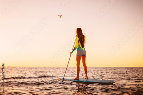 Young woman Stand Up Paddle Surfing and drone copter with beautiful sunset colors