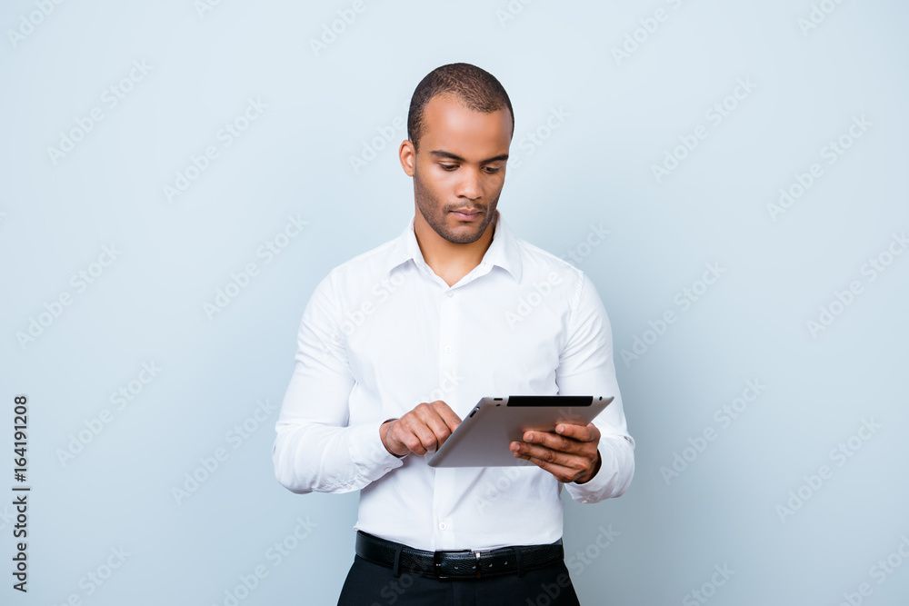 Successful ponder young african guy is browsing at his tablet, standing on the pure background, being pensive and severe Successful ponder young african guy is browsing at his tablet, standing on the pure background, being pensive and severe