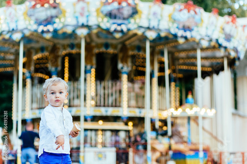 Funny toddler boy in white shirt and blue pants eating an ice cream in front of the carousel