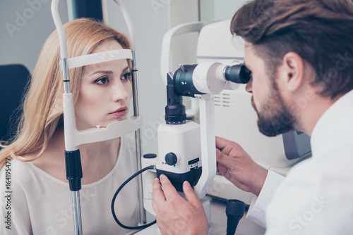 Concentrated brunet bearded optician with non contact tonometer is checking blond`s lady patient intraocular pressure at eye clinic. Health care, medicine, eye sight and technology concept photo