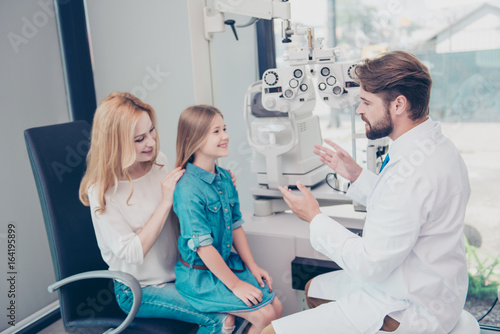 Optometrist consultation. Blond mom with smiling joyful girl are talking to brunet bearded male doctor optician in a hospital