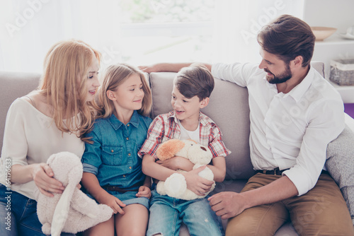 Fun time together. Happy family of four is listening to small boy kid telling about his adventures, all relatives look at him, smiling, at home on a couch photo