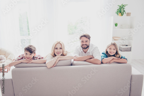 Happy family of four. Blond mom and small girl, brunet dad and little boy, lying on the beige sofa in a row, at home, all cheerful, smiling photo