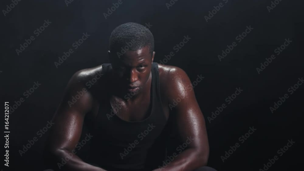 Zooming shot of a muscular black man resting in between workouts on a foggy dark background Zooming shot of a muscular black man resting in between workouts on a foggy dark background