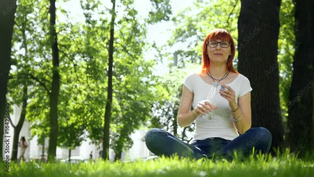 The red-haired girl drinking water sitting on grass in park The red-haired girl drinking water sitting on grass in park
