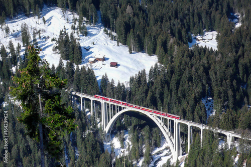 horizontal view of a red train crossing a bridge over a deep gorge in the Swiss Alps near Arosa and Chur in winter