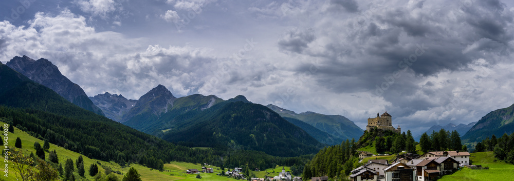 panorama view ofmountain peaks and a valley in the Swiss Alps with a small village and castle in the foreground underneath h a cloudy sky