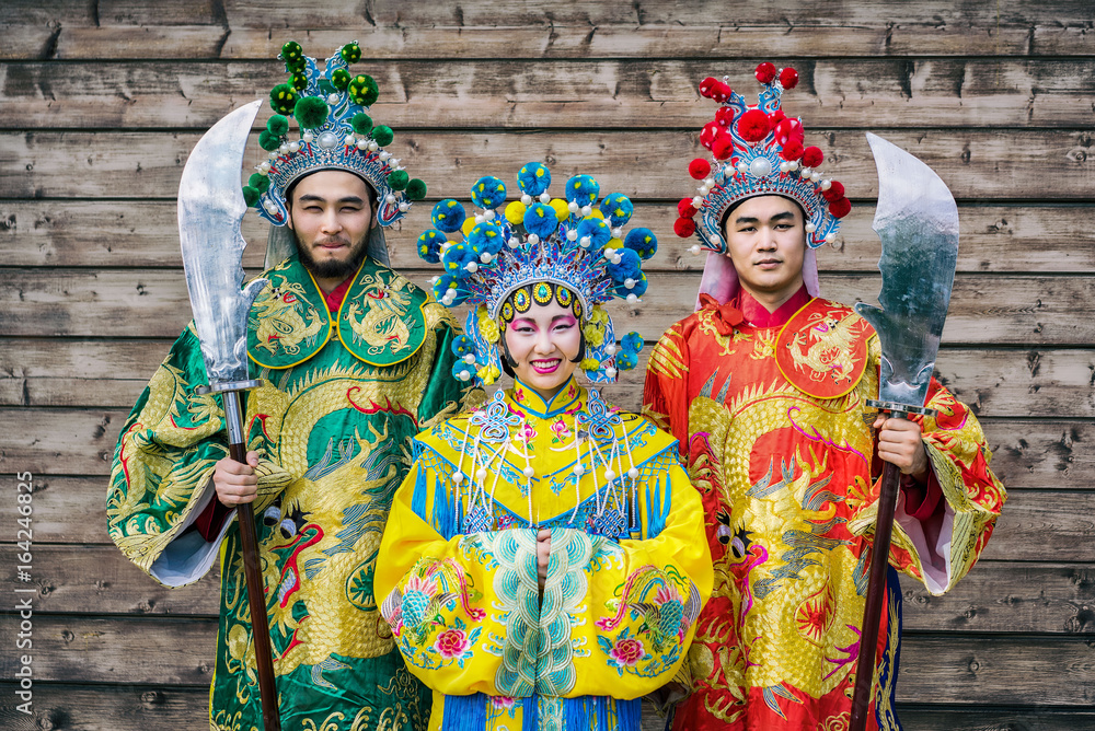 group of three people chinese men and woman in traditional costumes with swords greeting guests against wooden wall