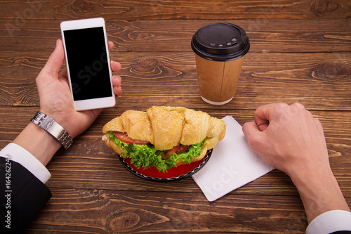 Croissant sandwich on a saucer, mobile phone in hand.Coffee on the table.