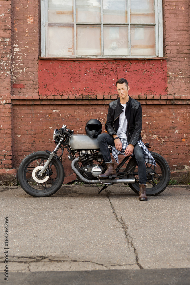 Handsome rider biker guy in black leather jacket, jeans and boots sit on  classic style cafe racer motorbike. Bike custom made in vintage garage.  Brutal fun urban lifestyle. Outdoor portrait. Stock Photo