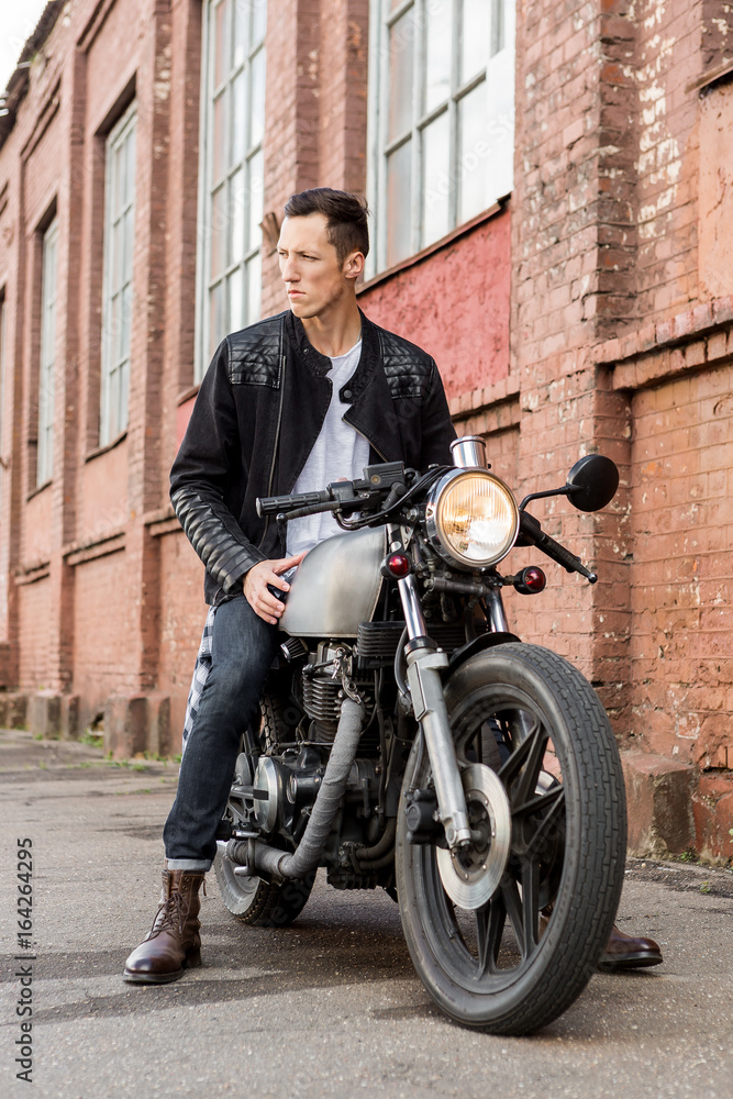 Handsome rider biker guy in leather jacket sit on classic style cafe racer motorcycle and look to the side. Bike custom made in vintage garage. Brutal fun urban lifestyle. Outdoor portrait.