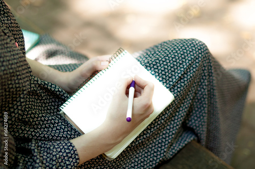 Legs of a  woman in long dress, on a bench in the park. Girl Write notes to notepad in a park outdoors Close-up. Point of view.