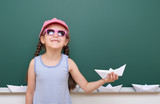 Schoolgirl with paper boat play near a blackboard, empty space, education concept