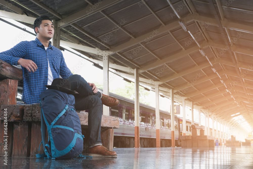 asian man with backpack sitting on platform at train station. backpacker or traveler waiting for train. journey, trip, travel concept